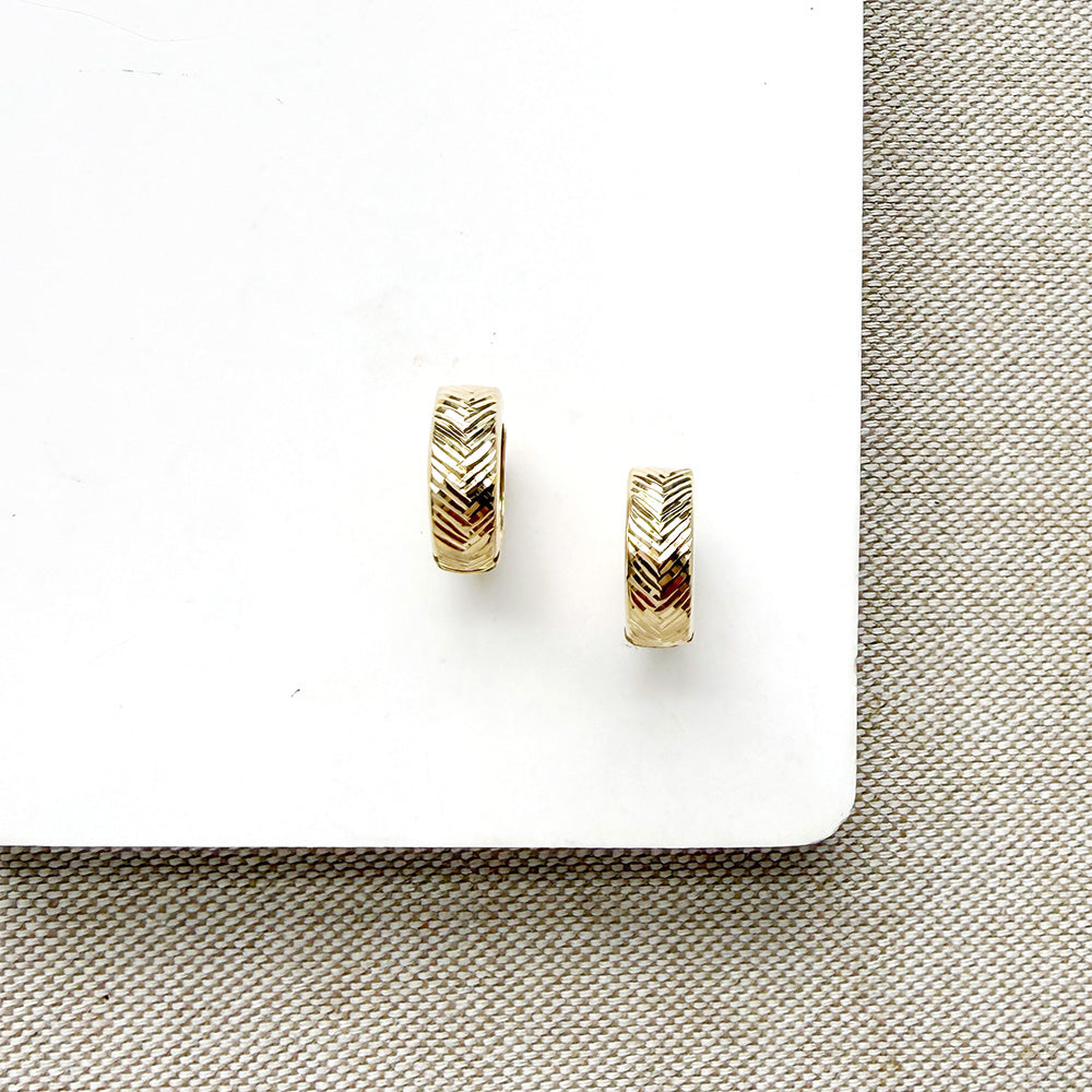 A pair of small gold huggie hoop earrings with a textured chevron pattern, displayed on a white card over a beige woven fabric background.