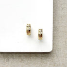 A pair of small gold huggie hoop earrings with a textured chevron pattern, displayed on a white card over a beige woven fabric background.