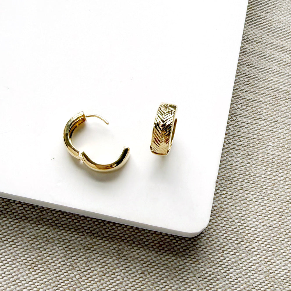 A pair of small gold hoop earrings, one shown from the side with a curved open-back design and the other from the front displaying a textured chevron pattern, resting on a white rectangular block over a woven beige surface.