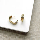 A pair of small gold hoop earrings, one shown from the side with a curved open-back design and the other from the front displaying a textured chevron pattern, resting on a white rectangular block over a woven beige surface.