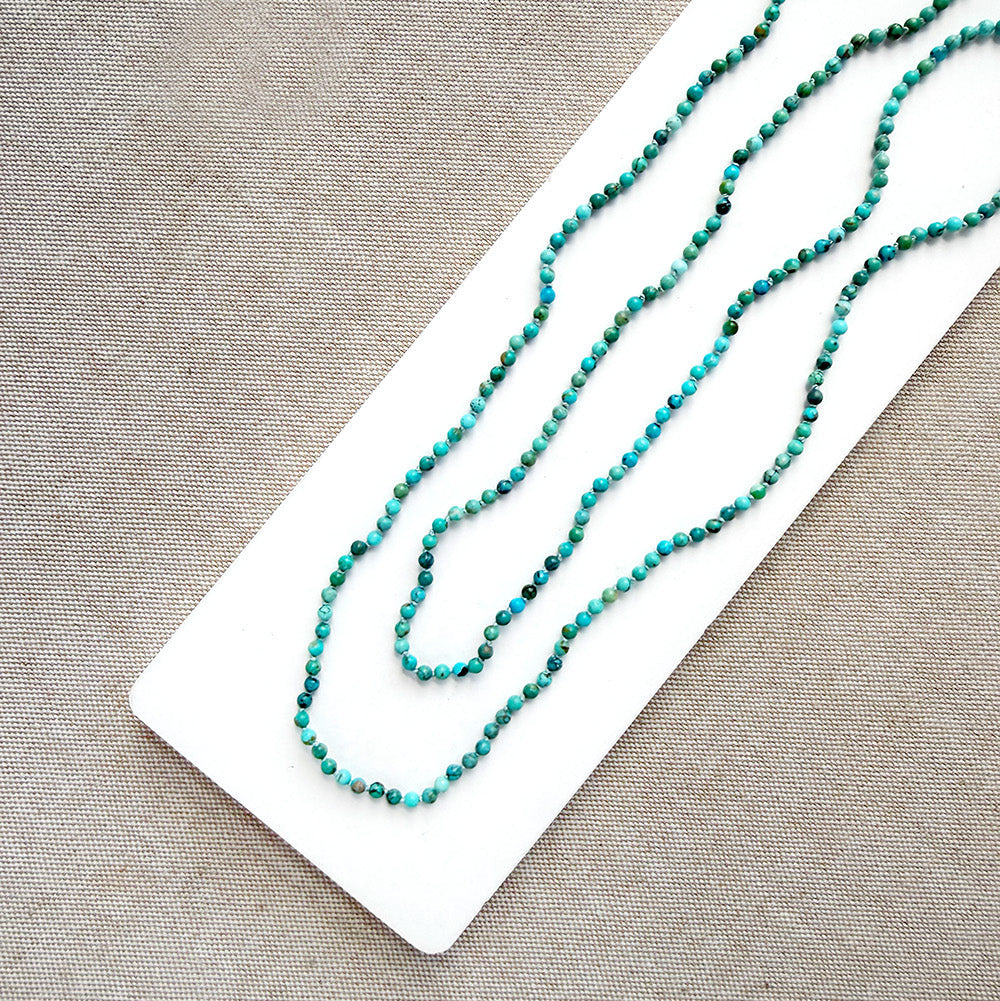 A long strand of small turquoise-blue beads draped diagonally across a white rectangular display card on a textured beige fabric background.