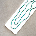 A long strand of small turquoise-blue beads draped diagonally across a white rectangular display card on a textured beige fabric background.