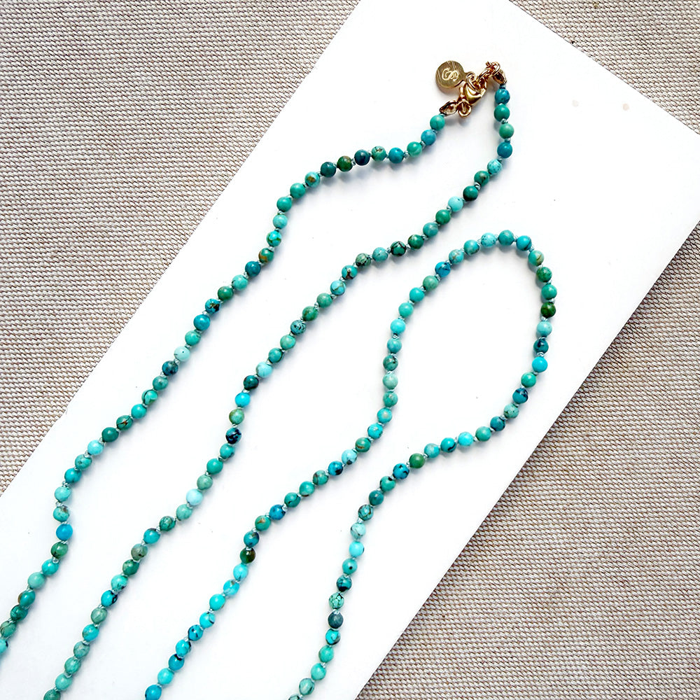 A beaded necklace made of small turquoise and green beads with a gold clasp, displayed on a white rectangular card over a textured beige fabric background.