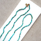 A beaded necklace made of small turquoise and green beads with a gold clasp, displayed on a white rectangular card over a textured beige fabric background.