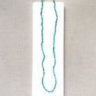 A long beaded necklace made of small turquoise-blue beads with a few dark accents, displayed on a white rectangular card against a textured light gray fabric background.