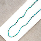 A beaded strand necklace made of small turquoise-blue and green marbled beads, laid diagonally on a white rectangular card over a beige woven fabric background.