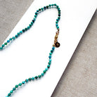 A beaded necklace made of small turquoise and green beads with a gold clasp and a small round charm, laid on a white card over a textured gray fabric background.