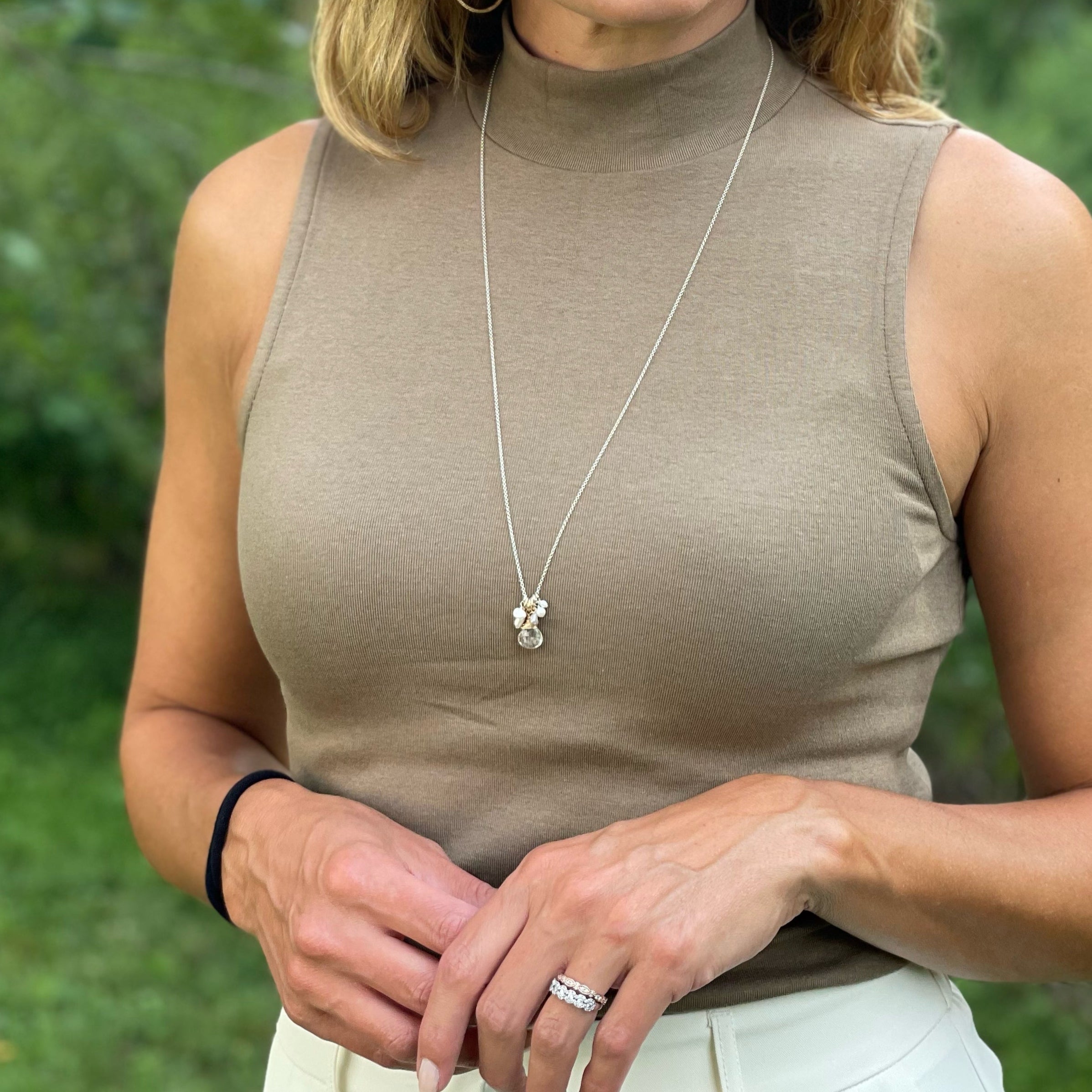 Woman wearing a beige sleeveless top with a necklace and ring, standing outdoors with greenery in the background.