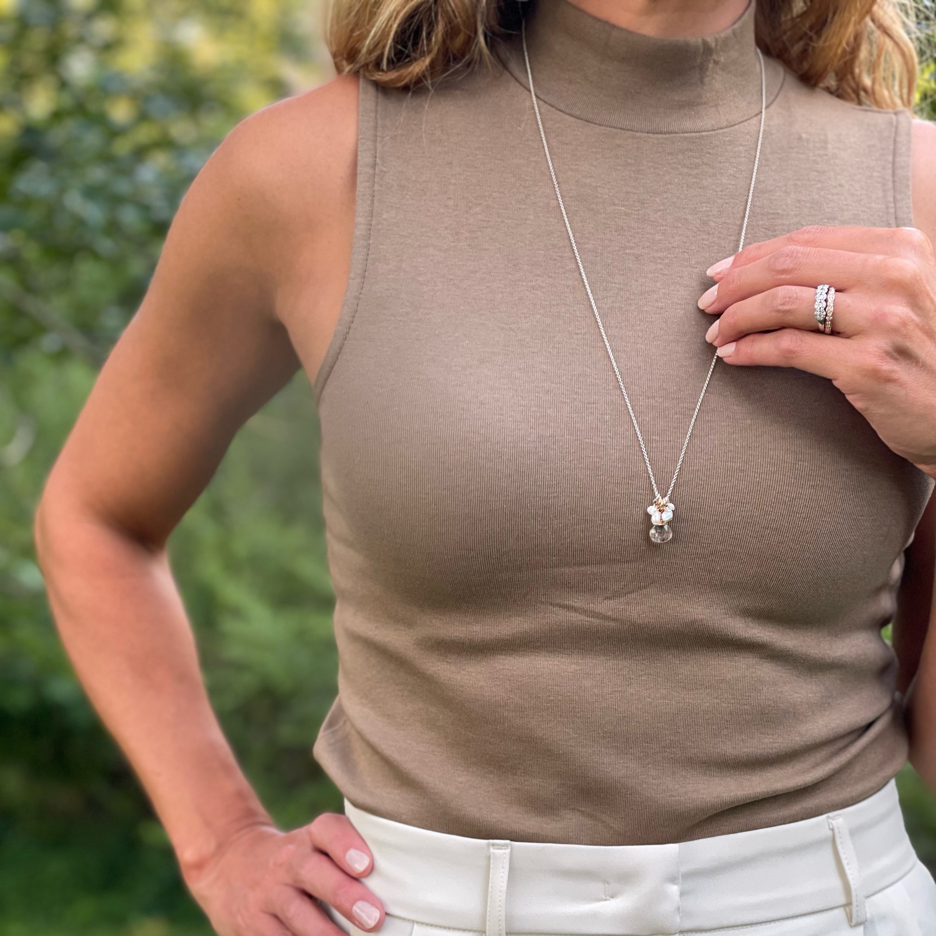 Person wearing a beige sleeveless top and white pants with a necklace outdoors.