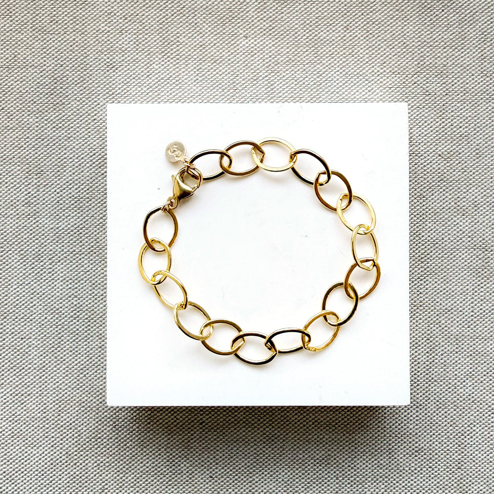 A gold chain link bracelet with oval links, displayed on a white square card over a textured gray fabric background.