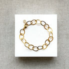 A gold chain link bracelet with oval links, displayed on a white square card over a textured gray fabric background.