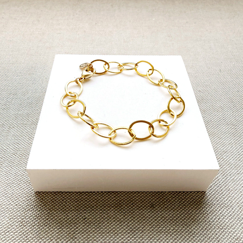 A gold chain link bracelet with oval links, displayed on a white square card over a textured gray fabric background.