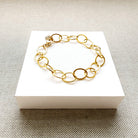 A gold chain link bracelet with oval links, displayed on a white square card over a textured gray fabric background.