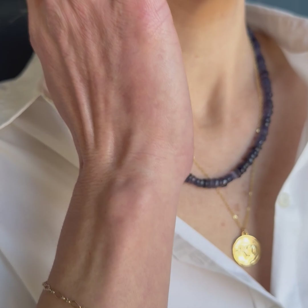 Close-up of a person wearing two necklaces, one with iolite gemstones and the other with a gold coin pendant, on a white shirt.