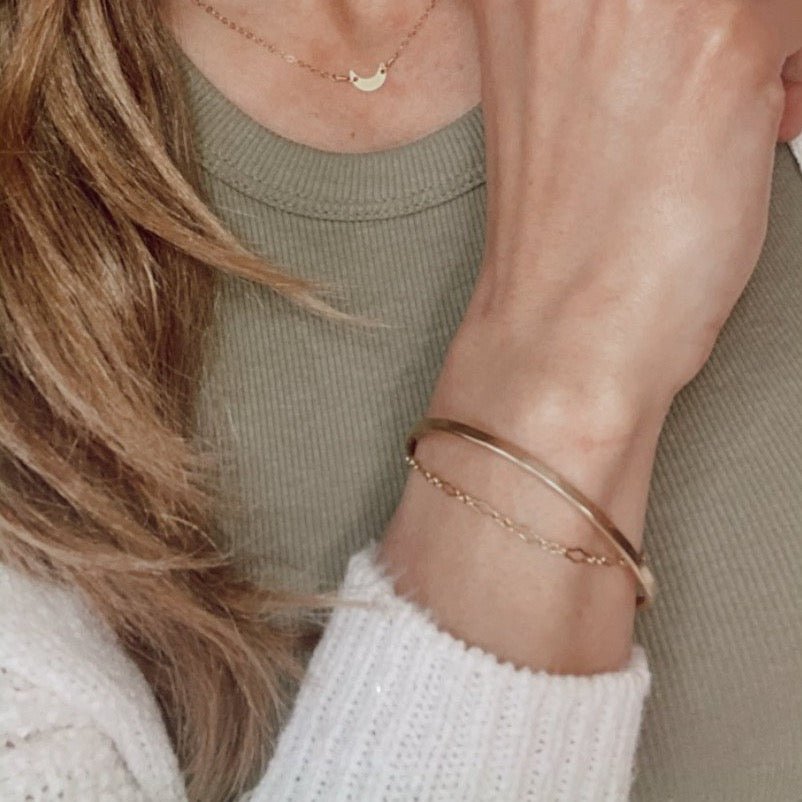 Woman wearing a green shirt and white sweater with gold textured bangle bracelet with the words "always with you" stamped on the inside, gold chain bracelet and gold moon necklace. Always with You Bangle by Sarah Cornwell Jewelry