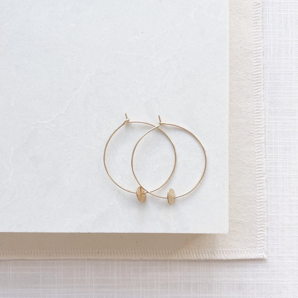 Gold Amalee Earrings by Sarah Cornwell Jewelry. Classic 1.5 inch gold hoop earrings with a small gold textured disc hanging from the bottom on a white background.