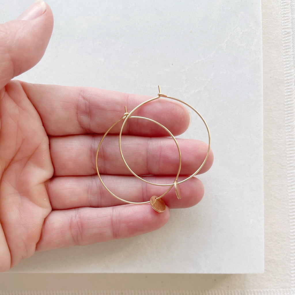 Gold Amalee Earrings by Sarah Cornwell Jewelry. Hand holding classic 1.5 inch gold hoop earrings with a small gold textured disc hanging from the bottom on a white background.