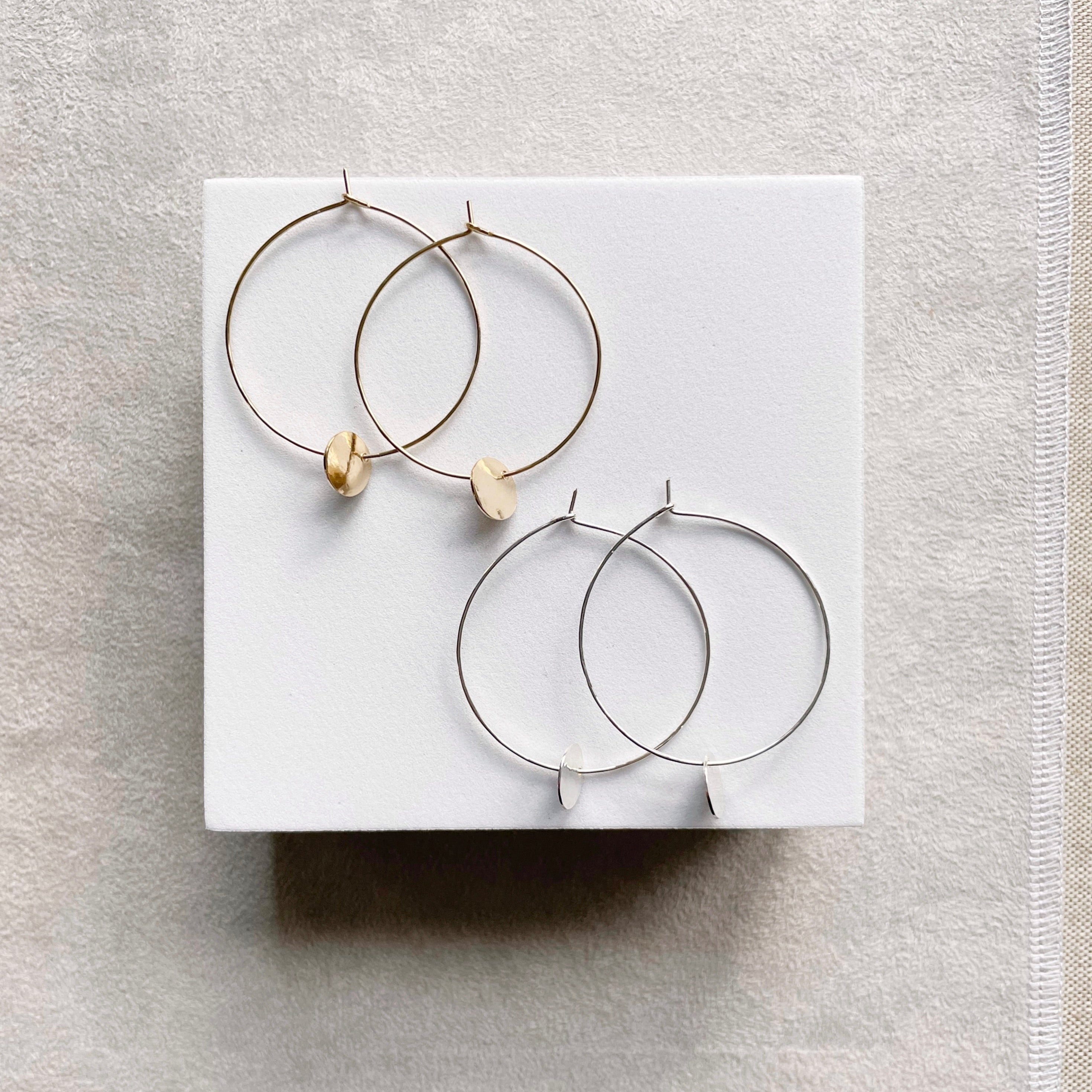 Amalee Earrings by Sarah Cornwell Jewelry. 2 pairs of gold and silver classic 1.5 inch gold hoop earrings with small gold textured discs hanging from the bottoms on a white background.