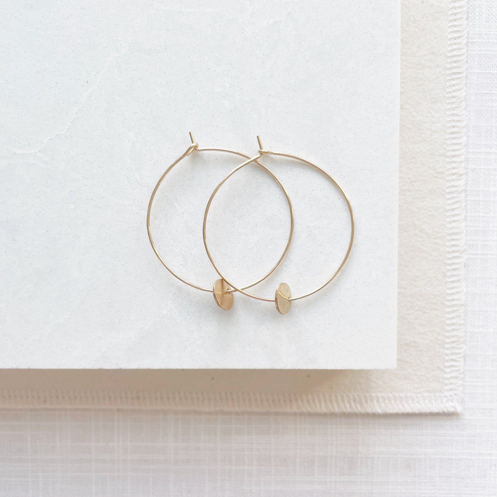 Gold Amalee Earrings by Sarah Cornwell Jewelry. Classic 1.5 inch gold hoop earrings with a small gold textured disc hanging from the bottom on a white background.