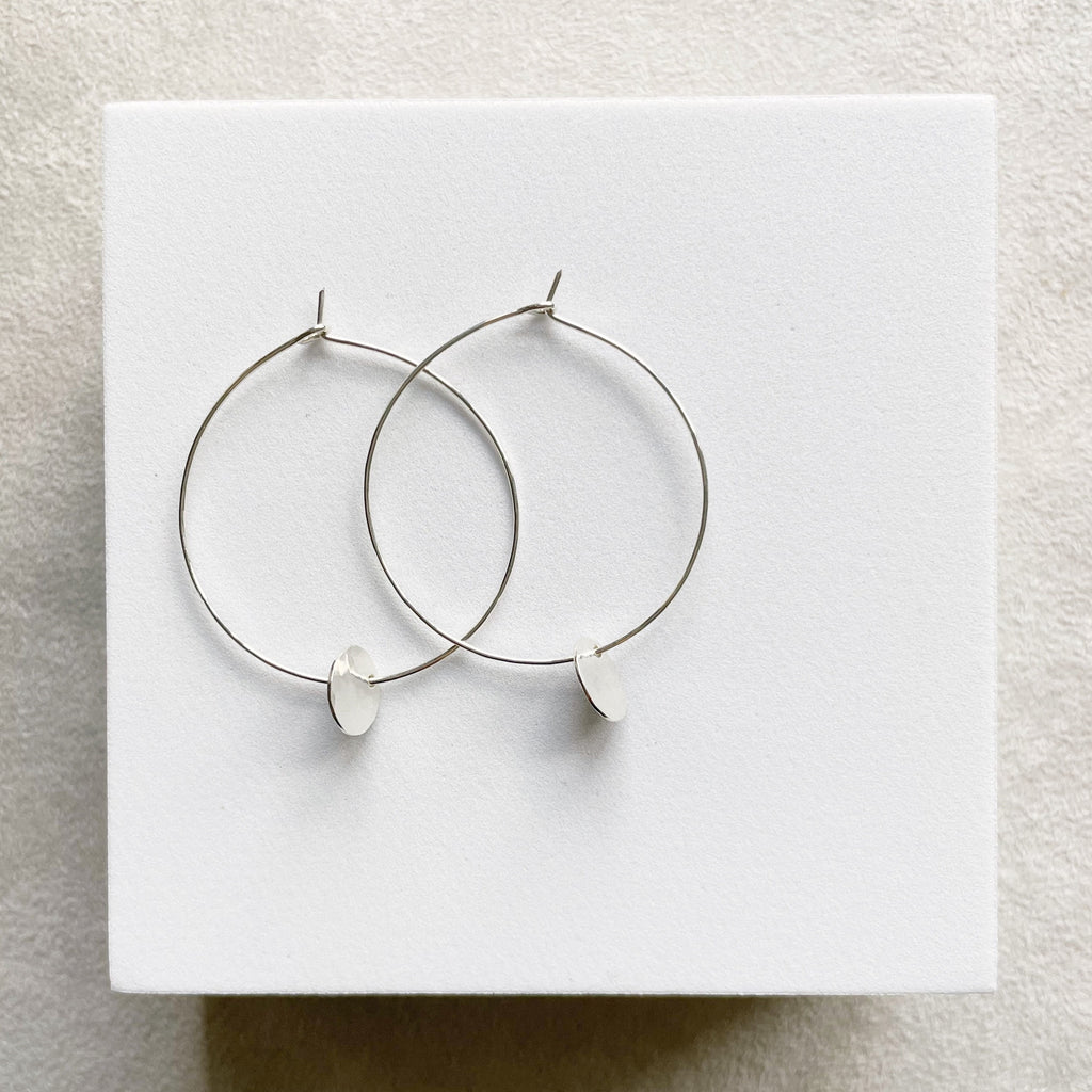 Silver Amalee Earrings by Sarah Cornwell Jewelry. Classic 1.5 inch silver hoop earrings with a small silver textured disc hanging from the bottom on a white background.