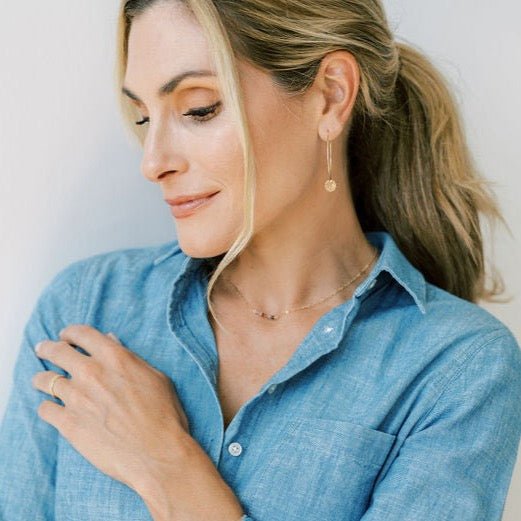 Woman with blonde hair in a ponytail wearing denim button down with dainty gold 16 inch gemstone necklace with sapphires in warm shades on a straight bar and gold textured disc earrings. Andra necklace by Sarah Cornwell Jewelry