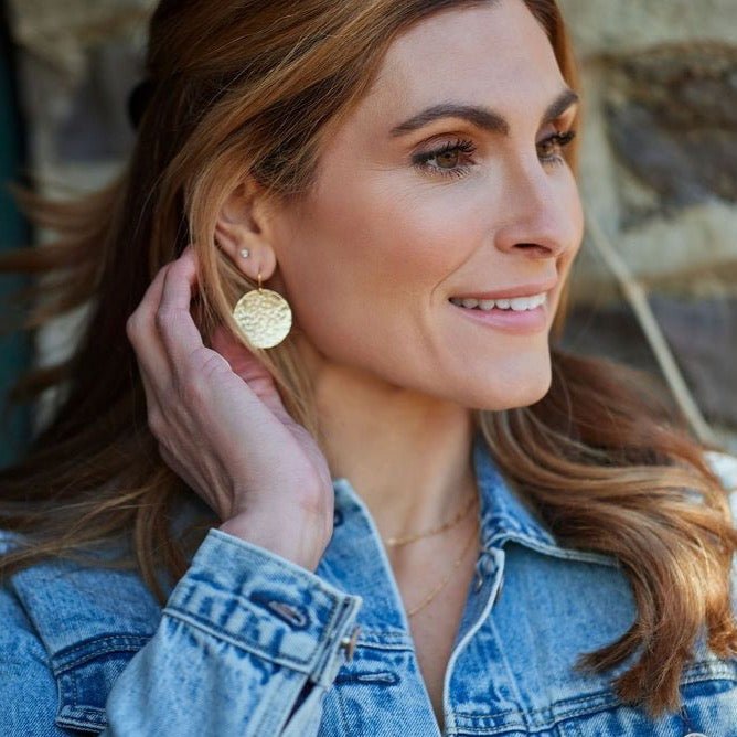 Gold Buffay Earrings by Sarah Cornwell Jewelry. Blonde woman wearing denim jacket, lightweight, shimmery, textured 1 inch gold disc earrings stacked with gold studs and gold layered necklaces.