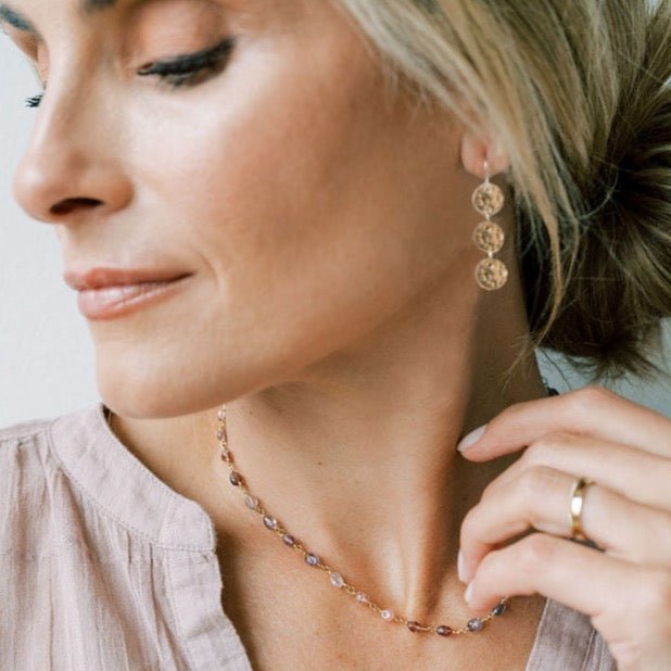 Side view of woman's face and neckline with blonde hair up wearing a taupe shirt with 17 inch gold gemstone necklace with full strand of wire wrapped gradient colored red, purple and pink spinel gemstones and gold textured disc drop earrings with 3 discs and a gold ring. Cain Necklace by Sarah Cornwell Jewelry