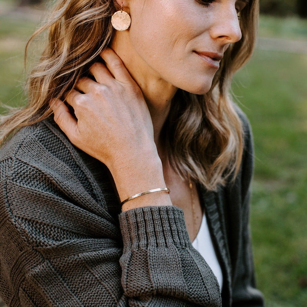 Side view of woman with long blonde hair wearing gray sweater with gold textured bangle bracelet and gold textured disc statement earrings. Caroline Bangle by Sarah Cornwell Jewelry