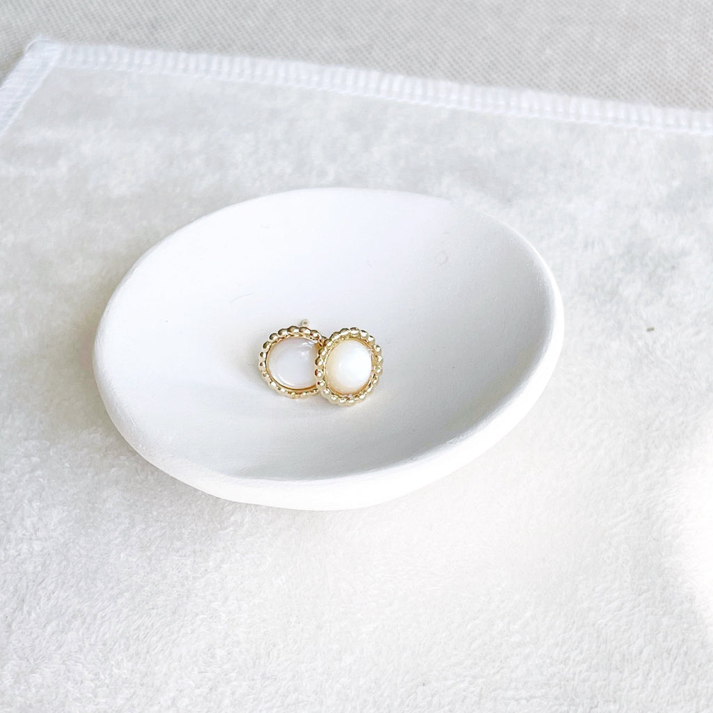 Pair of 6mm gold and mother of pearl stud earrings with a beaded bezel setting in a white dish. Chantilly Studs by Sarah Cornwell Jewelry