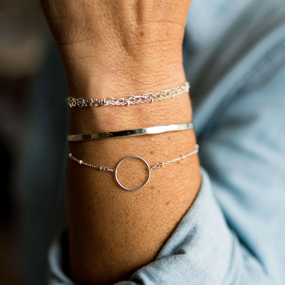 Woman's wrist wearing blue shirt with silver textured bangle bracelet with initials stamped on both ends and silver circle bracelet and silver multi chain bracelet. Charlie Bangle by Sarah Cornwell Jewelry