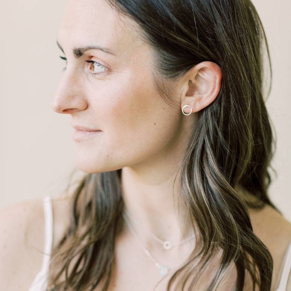 Side view of woman with long brown hair wearing a white top with gold simple circle stud earrings and layered gemstone necklaces. Circle Studs by Sarah Cornwell Jewelry