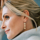 Close up side view of a woman with blonde hair wearing a gray sweater with gold 1.5 inch drop earrings with 3 same sized textured discs and gold huggie hoops in her second piercing. Cole Earrings by Sarah Cornwell Jewelry