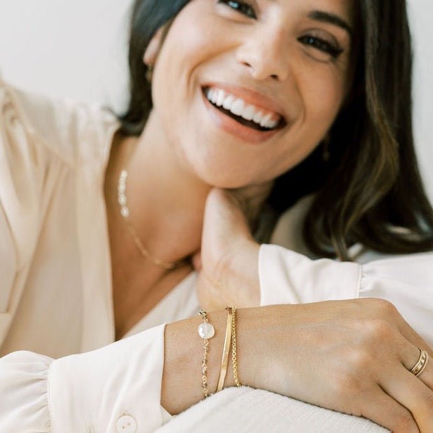 Woman with long dark hair wearing cream blouse with gold rolo style chain bracelet, gold chain and white chalcedony bracelet, gold bangle, gold chain necklace and gold ring stack. Danny Bracelet by Sarah Cornwell Jewelry