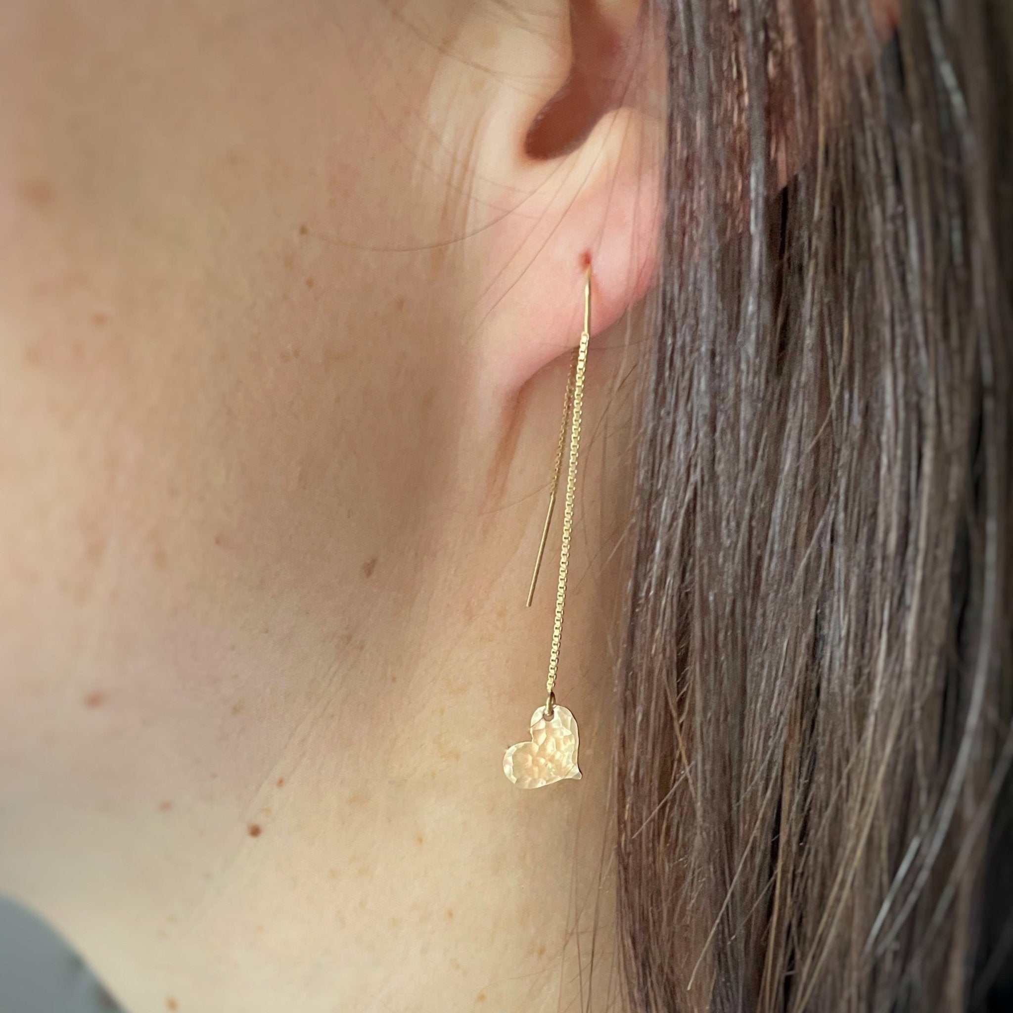 Close up side view of woman with dark hair wearing gold chain threader earrings with a 2.25 inch drop and a tiny textured gold heart at the bottom. Darling Earrings by Sarah Cornwell Jewelry