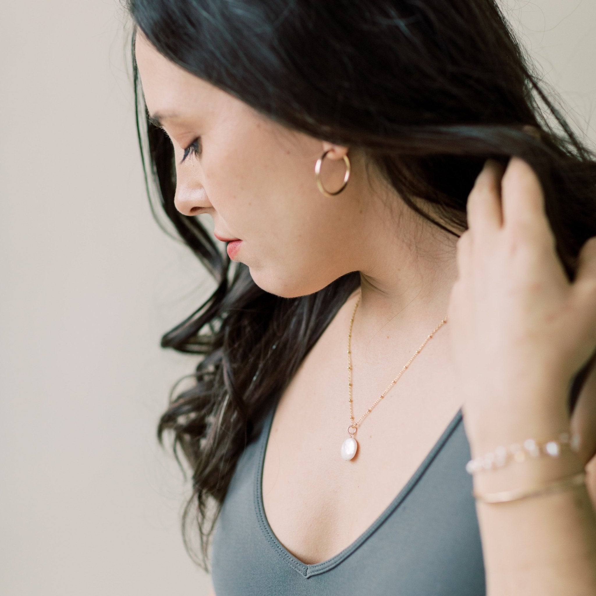 Side view of woman with long dark hair wearing a gray shirt with gold and coin pearl long statement necklace on a link ball chain and layered bracelets and gold hoop earrings. Deene Necklace by Sarah Cornwell Jewelry