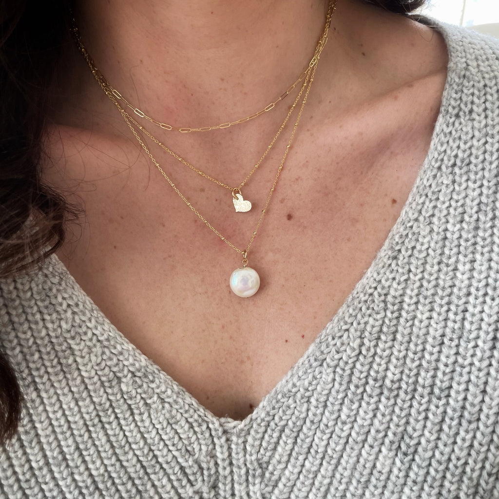 Woman's neckline wearing a gray sweater with gold and coin pearl long statement necklace on a link ball chain with gold chain and gold heart layered necklaces and gold stud earrings. Deene Necklace by Sarah Cornwell Jewelry