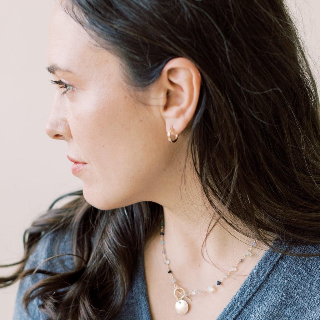 Gold Demi Hoops by Sarah Cornwell Jewelry. Dark haired woman wearing denim shirt with small, simple, everyday gold hoop earrings and gold textured disc and gemstone toggle necklace.