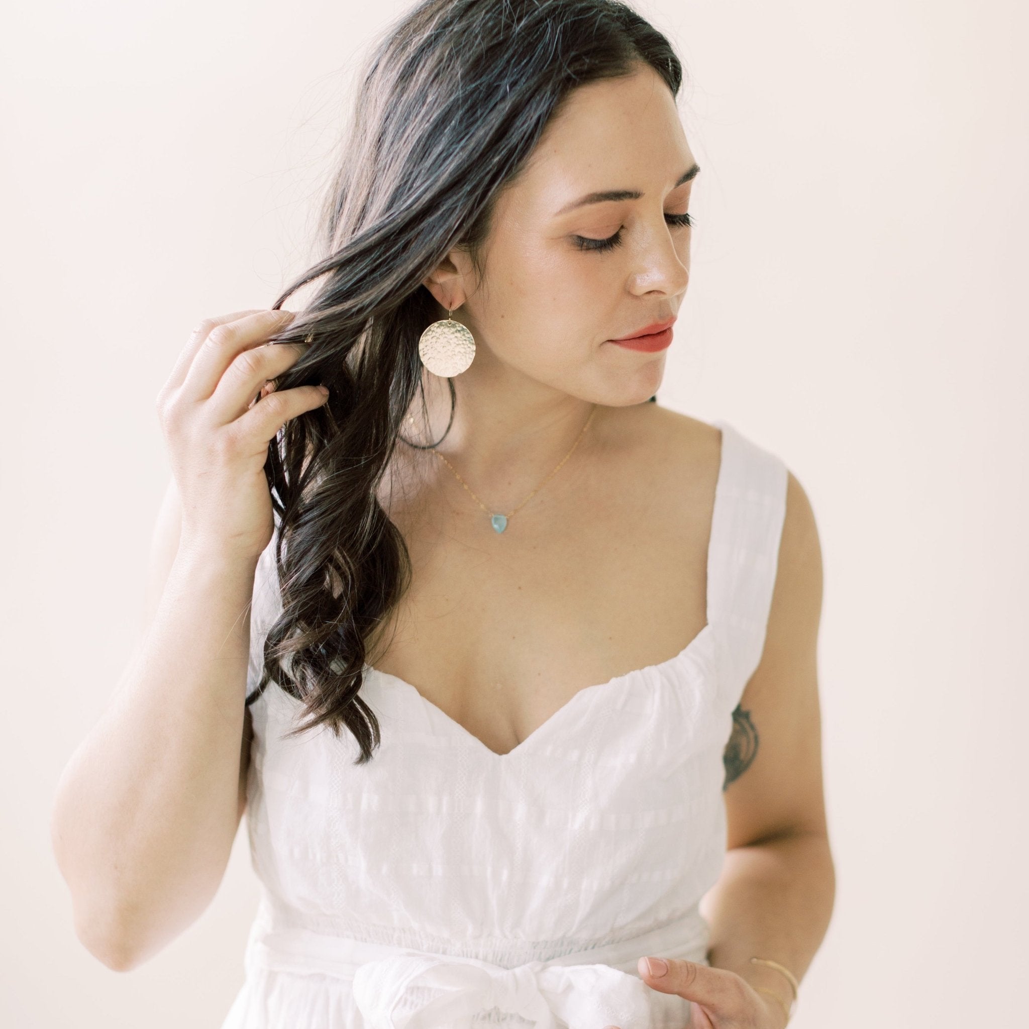 Woman with long black hair wearing white sleeveless top with 17 inch gold pendant necklace with teardrop shaped aqua chalcedony pendant and large gold textured disc statement earrings. Hampton Necklace by Sarah Cornwell Jewelry