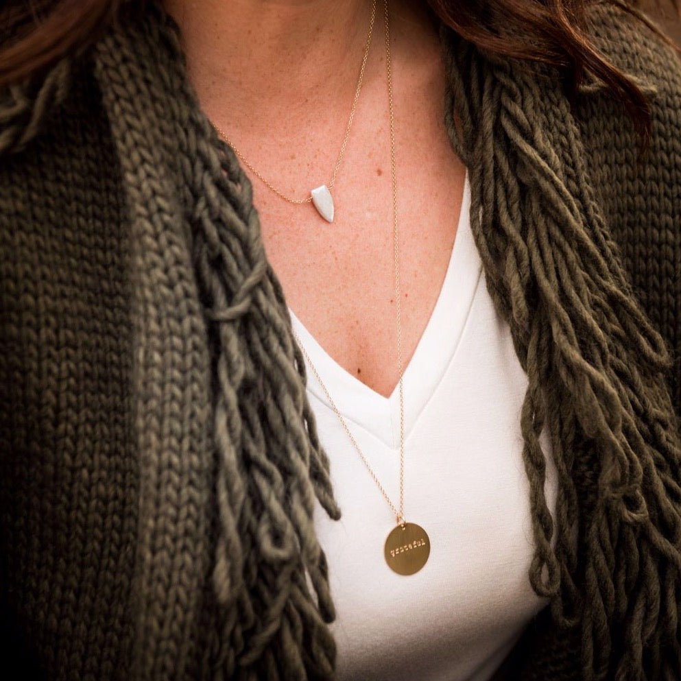 Woman's neckline wearing white v neck and green sweater with 28 inch gold necklace with 3/4 inch stamped disc with the word grateful layered with gemstone pendant necklace. Hana Necklace by Sarah Cornwell Jewelry