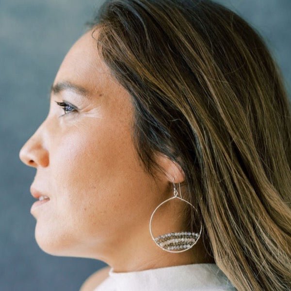 Woman with long dark hair wearing a white shirt with gold and gemstone statement 2 inch hoop earrings with rows of zircon, labradorite, and smokey topaz gemstones. Harper Earrings by Sarah Cornwell Jewelry