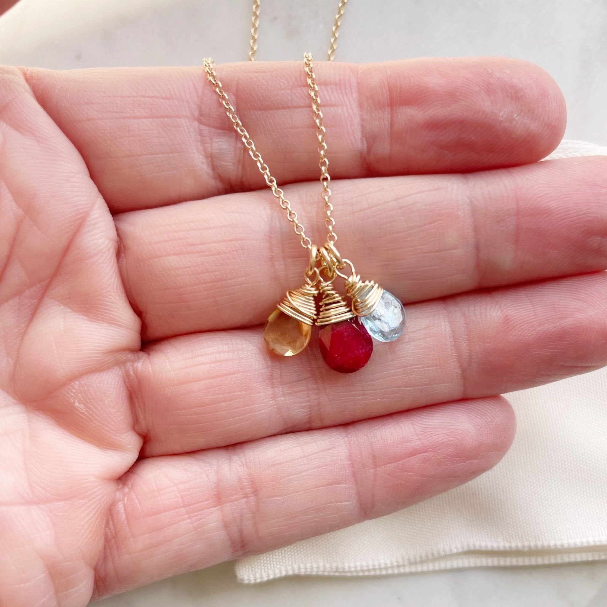 Hand holding gold wire wrapped gemstone birthstone necklace with yellow, red and light blue gemstone pendants. Heritage Necklace by Sarah Cornwell Jewelry