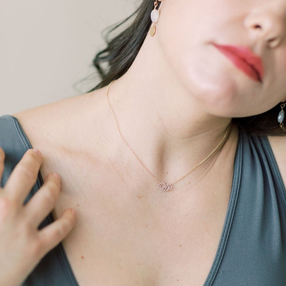 Woman's neckline with dark hair and red lipstick wearing gray tank top with gold necklace with 5 tiny gold rings and pearl and textured disc drop earrings. Honor Necklace by Sarah Cornwell Jewelry