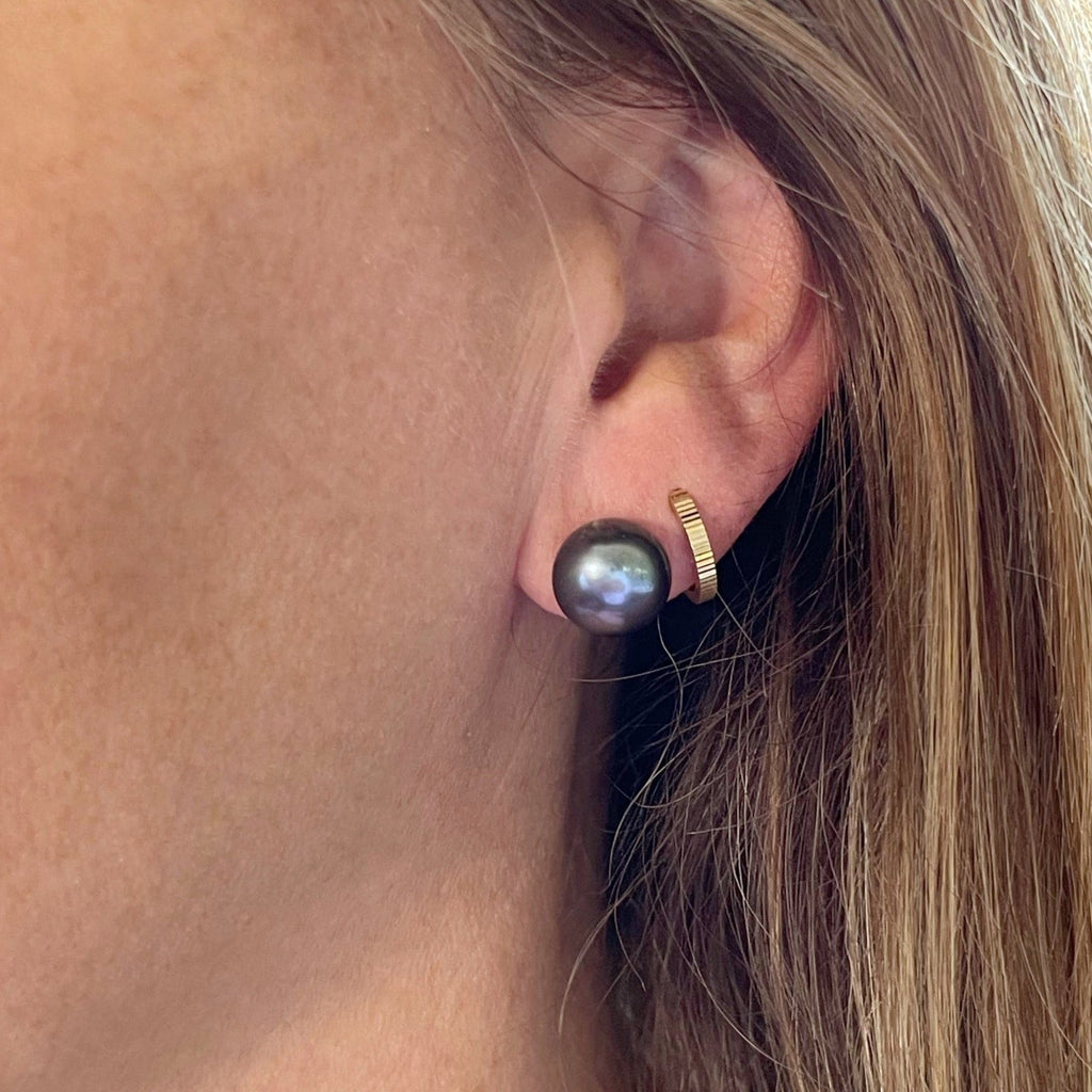 Close up side view of a woman wearing deep blue 11 mm freshwater pearl stud earrings in a silver setting stacked with gold huggie hoop earrings in the second piercing. Hudson Pearl Studs by Sarah Cornwell Jewelry