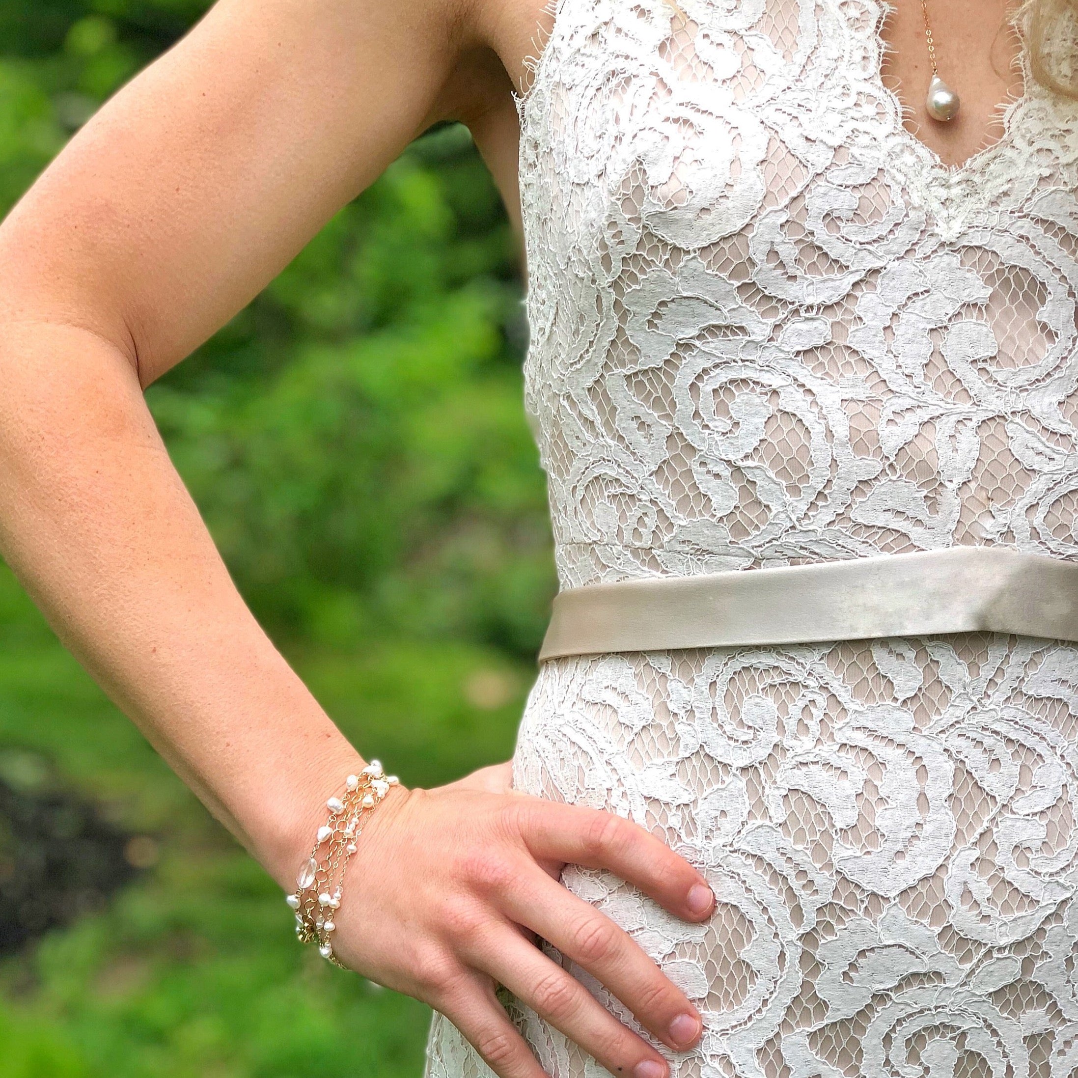Close up of woman's wrist and waist wearing  lace wedding dress with satin belt with gold double strand wire wrapped pearl bracelet and pearl drop necklace. Poppy Linen Bracelet by Sarah Cornwell Jewelry