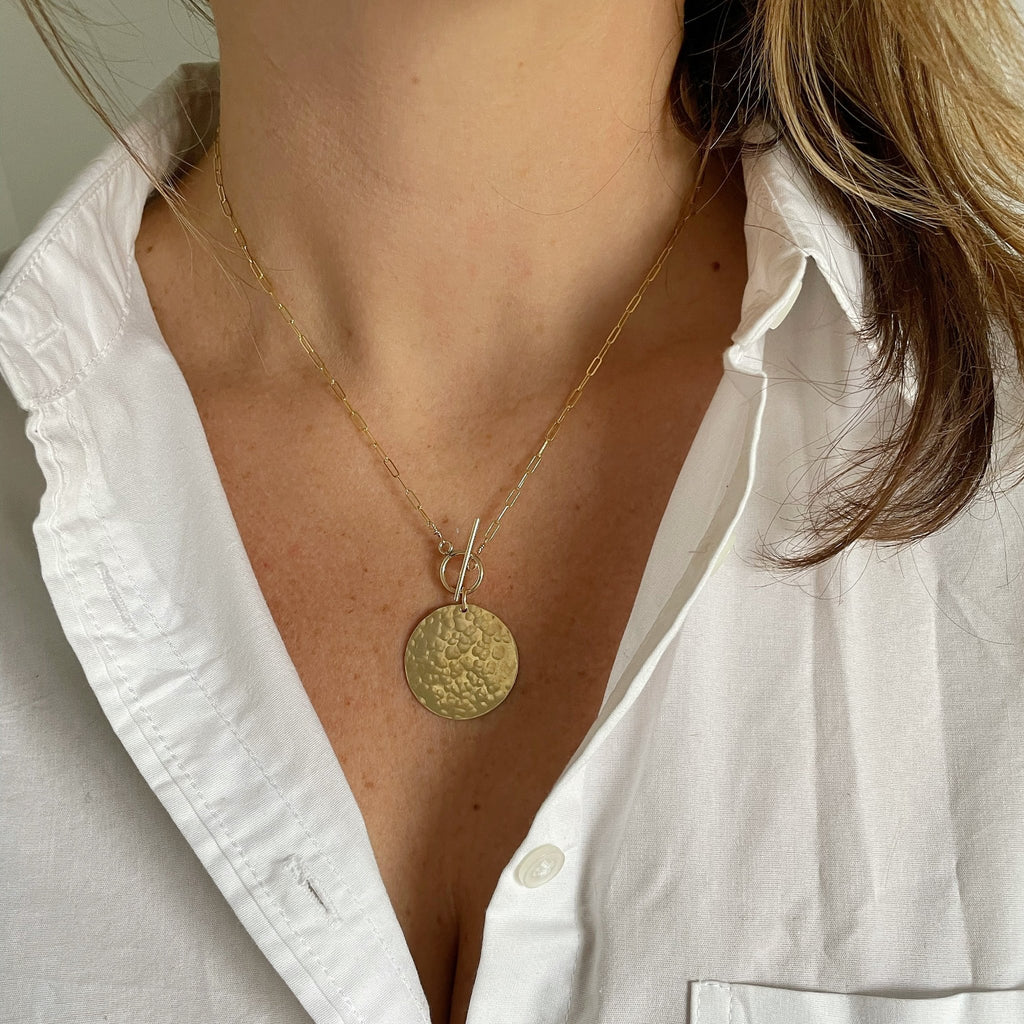Woman's neckline wearing white button down with gold statement necklace with large gold textured disc and front toggle. Josephine Necklace by Sarah Cornwell Jewelry