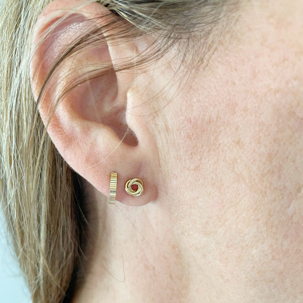 Woman with blonde hair wearing 6 mm gold love knot stud earrings and gold huggie hoop earrings. Love Knot Studs by Sarah Cornwell Jewelry