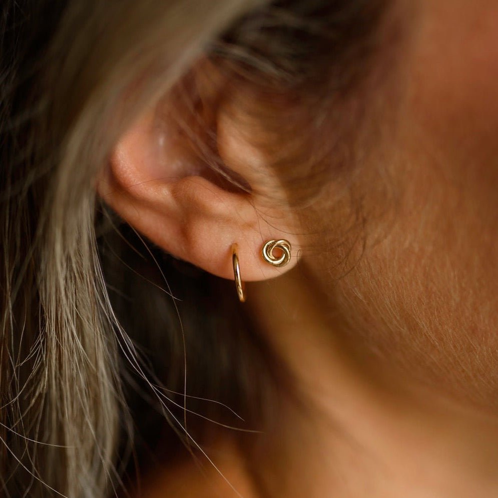 Woman with blonde hair wearing 6 mm gold love knot stud earrings and gold huggie hoop earrings. Love Knot Studs by Sarah Cornwell Jewelry