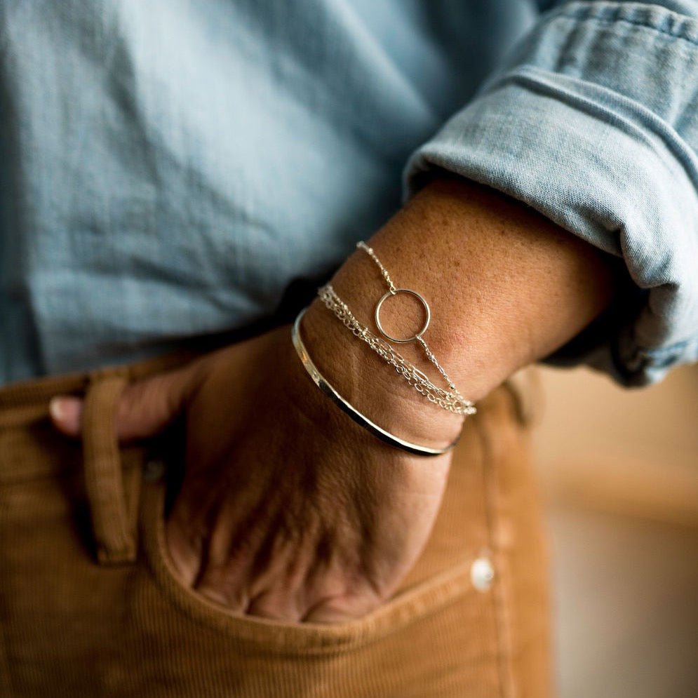 Close up of woman's wrist wearing blue shirt and tan corduroy pants with gold 3 strand bracelet with rectangle chain, link chain, and link ball chains, gold circle bracelet and gold bangle bracelet. Mina Bracelet by Sarah Cornwell Jewelry