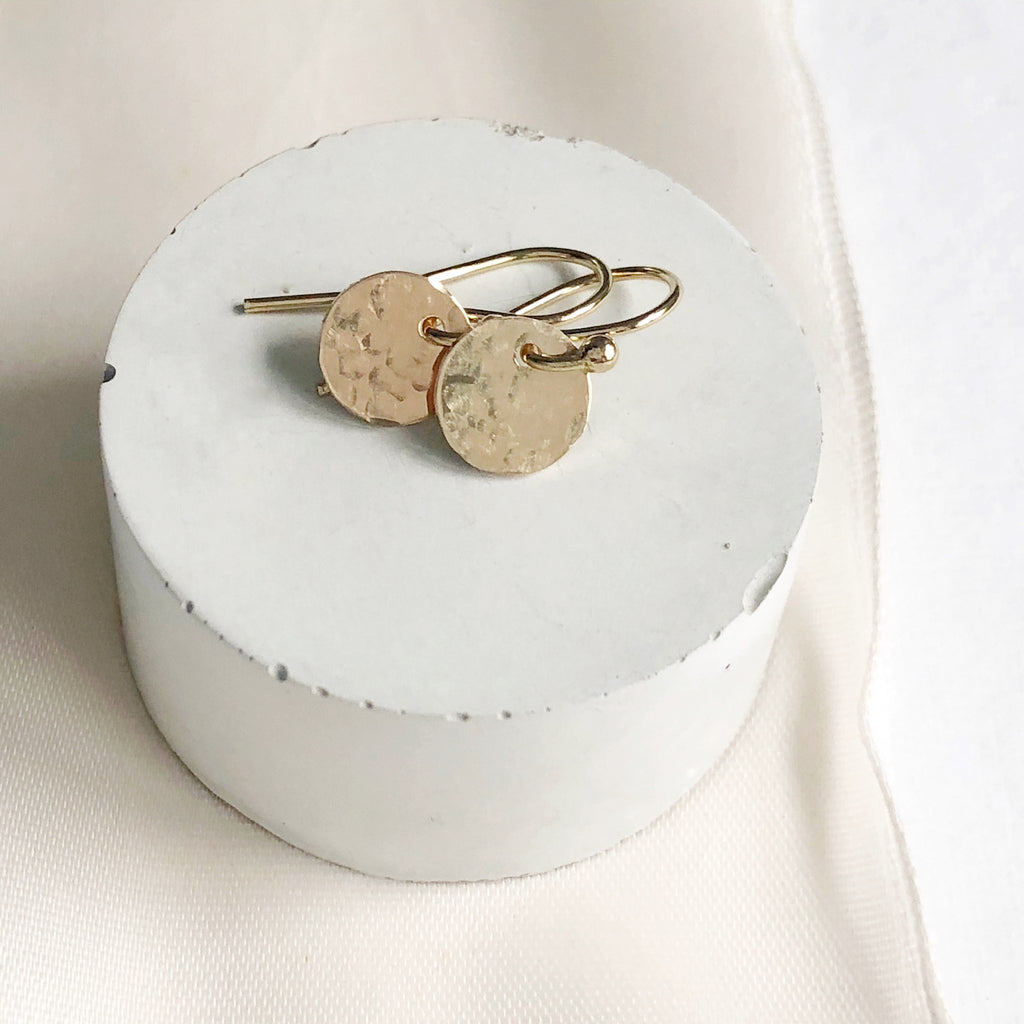 Gold Monica Earrings by Sarah Cornwell Jewelry. Shimmery, dainty gold 3/8 inch textured disc earrings on a round white riser.