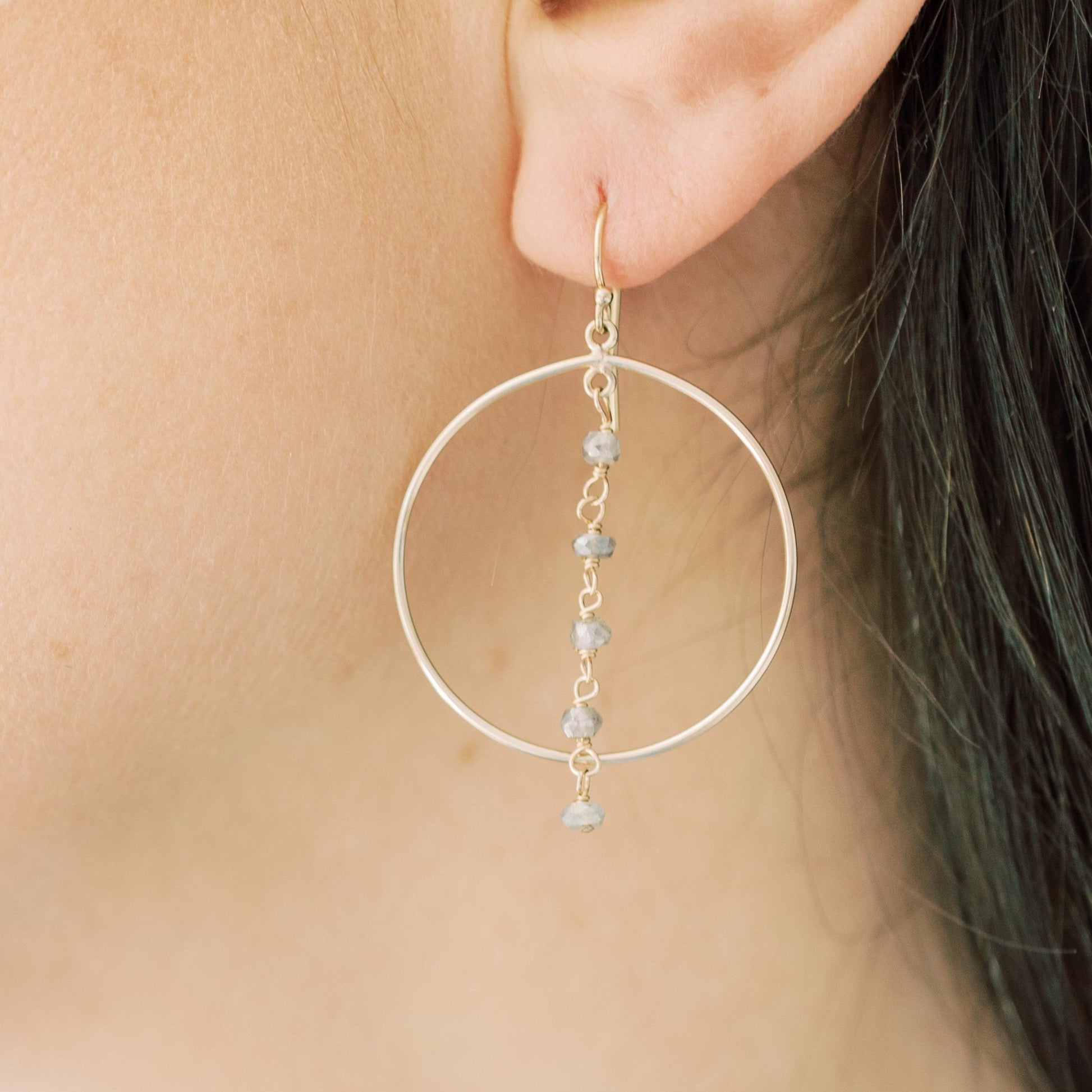 Gold Nantucket Earrings by Sarah Cornwell Jewelry. Woman's ear with sparkly statement gold hoop earrings with a strand of 5 wire wrapped labradorite gemstones down the middle.